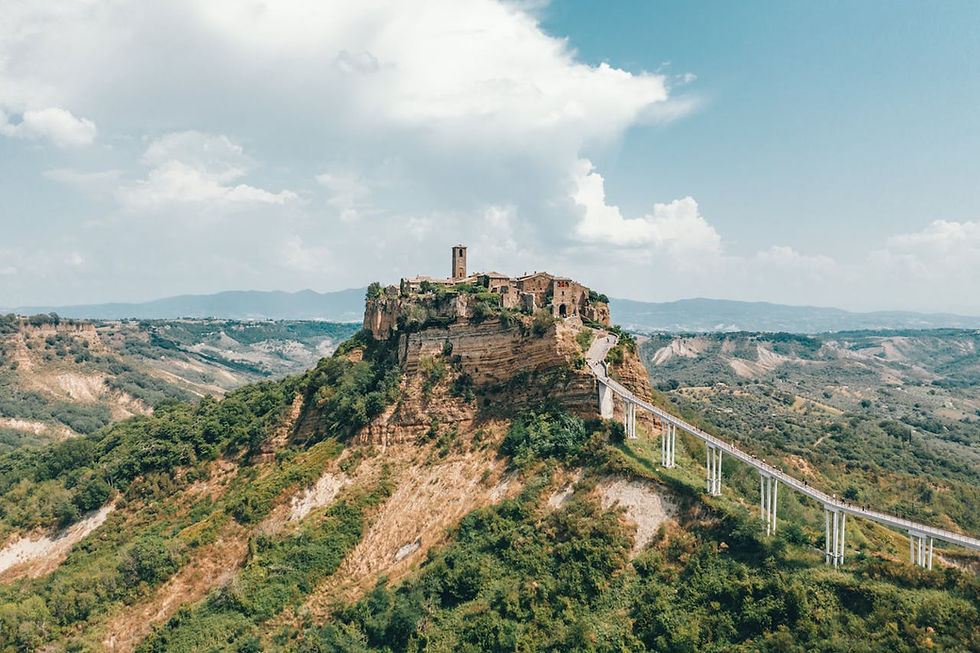 Civita di Bagnoregio, Italy with stone buildings, tower, and bridge set against lush green valleys and a bright blue sky with clouds. Peaceful atmosphere.