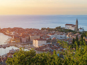 Scenic view of Piran, Slovenia, featuring red-roofed buildings, a church, marina with boats, and lush greenery in the foreground.
