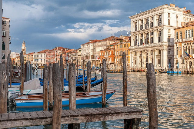 Canal scene in Venice with boats docked at wooden piers. Historic buildings in warm sunlight line the water. Cloudy sky casts a serene mood.