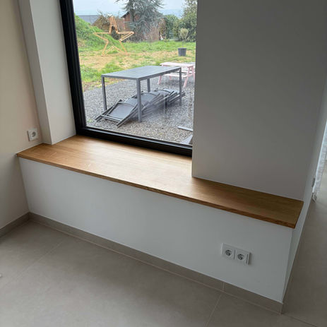 Maßgefertigte Fensterbank aus hellem Holz mit weißem Untergestell vor einem großen Fenster