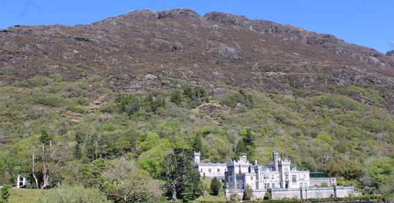 L'abbaye de Kylemore en pierre claire au bord de l'eau, dominée par une immense montagne rocheuse et verdoyante sous un ciel bleu.