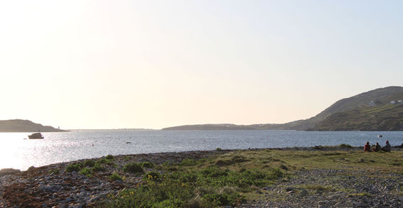 Rivage sauvage de Bay Beach à Clifden avec herbe, galets et un bateau au loin sur l'océan face aux collines d'Irlande.