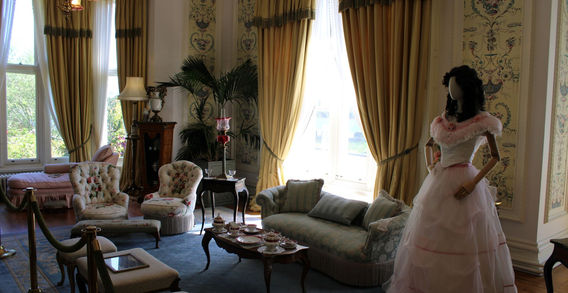 Un grand salon victorien avec des canapés, des fauteuils, des rideaux dorés et un mannequin en robe de bal blanche et rose à l'abbaye de Kylemore.