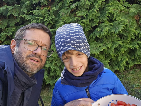 Egy szemüveges, szakállas férfi és egy láthatóan genetikai eltéréssel élő kisfiú mosolyognak a kamerába és egy paradicsomokkal teli zománcos tálat tartanak. Mögöttük hatalmas zöld tuják.