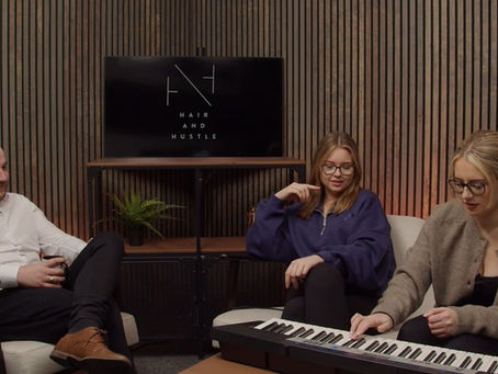 Three people in a wood-paneled room, one playing a keyboard. Relaxed mood. A screen reads "Hair and Hustle."