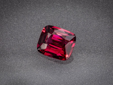 Faceted red gemstone on a gray textured surface, sparkling under light with no text visible.
