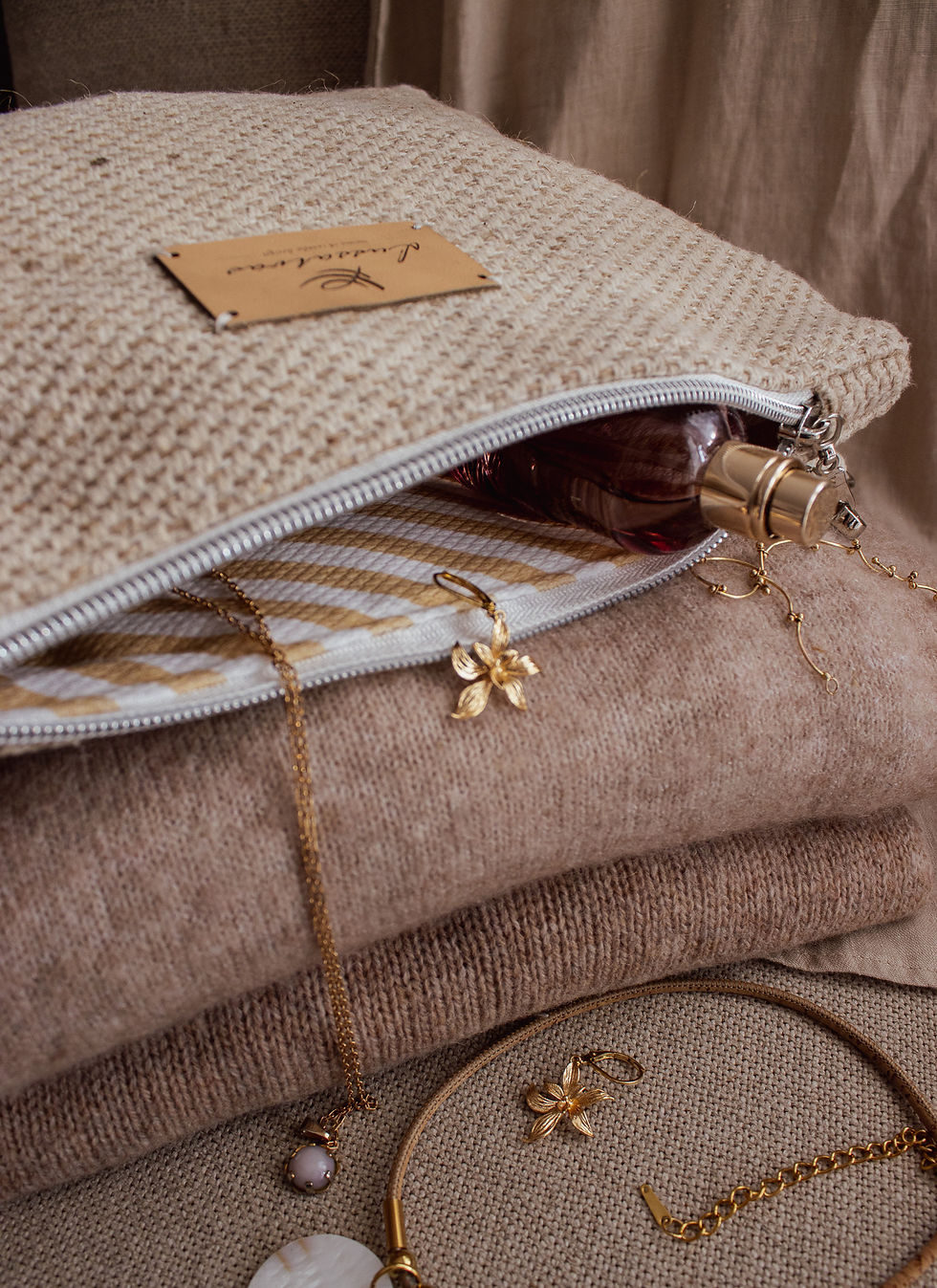 Pouch aus grobem Naturtextil mit hochwertigem Leder-Logo und Goldschmuck-Arrangement. Gestreiftes Futter