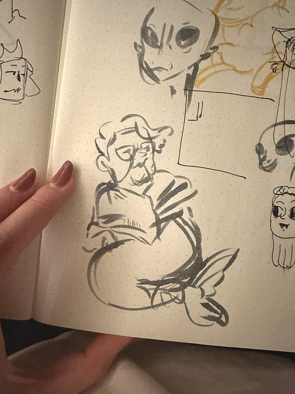 Notebook page with sketches of various faces, including an alien and a person with glasses. A hand with red nails holds the page.