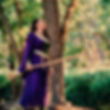 Woman in a flowing purple and green medieval-style dress holding a broomstick, standing in a sunlit forest and looking up with a joyful expression.