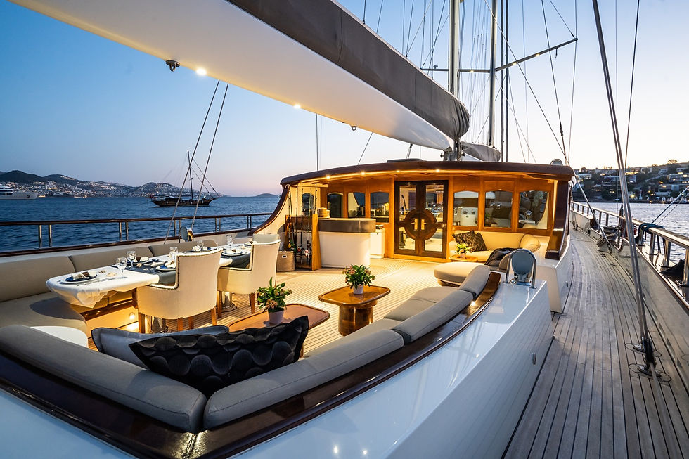 Luxurious yacht deck at sunset with dining table set for six, beige chairs, and potted plants. Calm sea and hilly shoreline in background.
