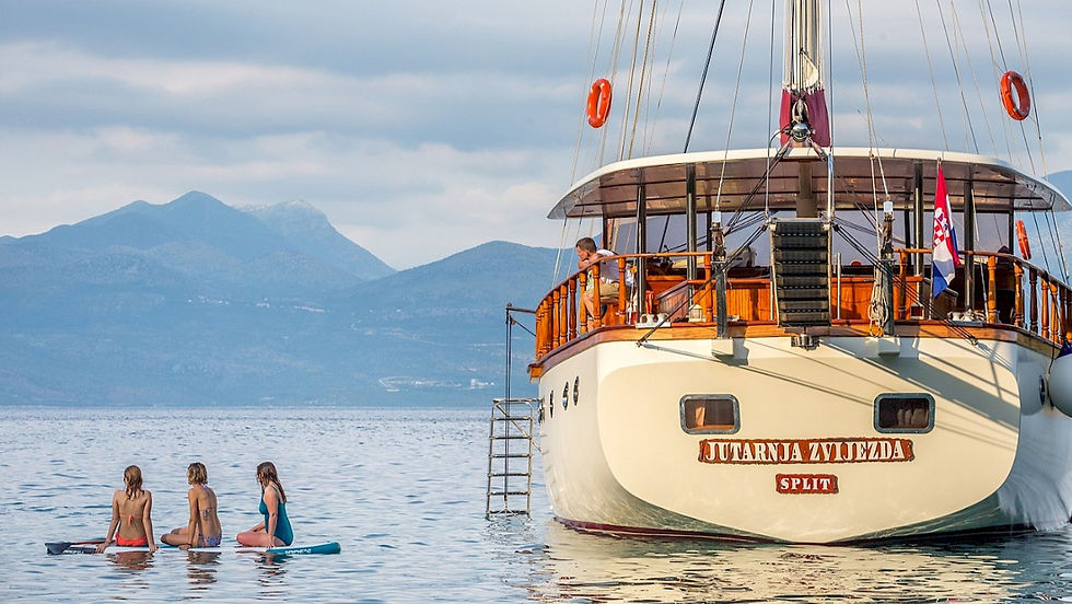 Three people on paddleboards in a calm sea, beside a wooden ship named "MORNING STAR," mountains in the backdrop, under a cloudy sky.