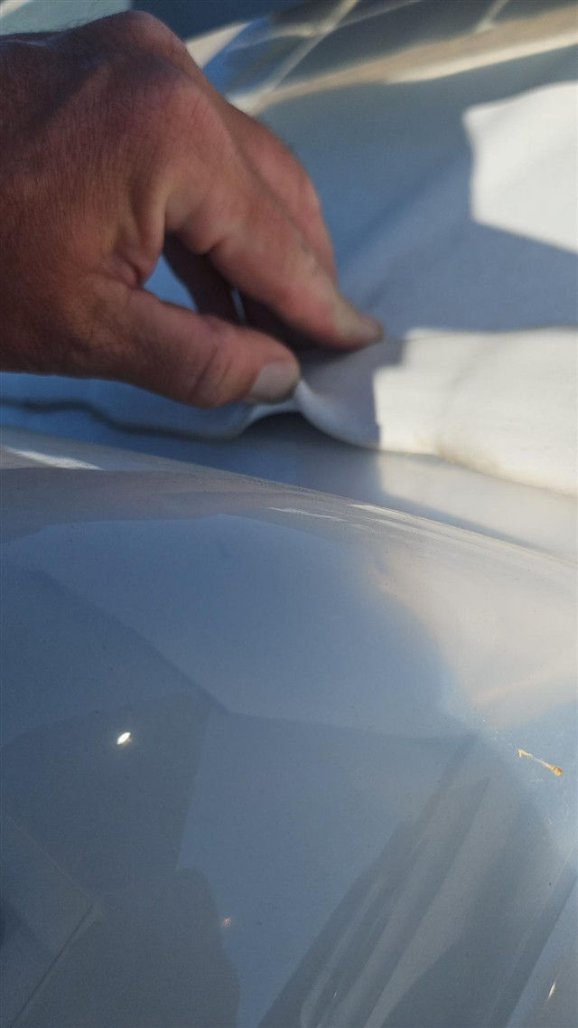 Hand pinching white material on a glossy blue car surface in bright sunlight, with strong reflections.