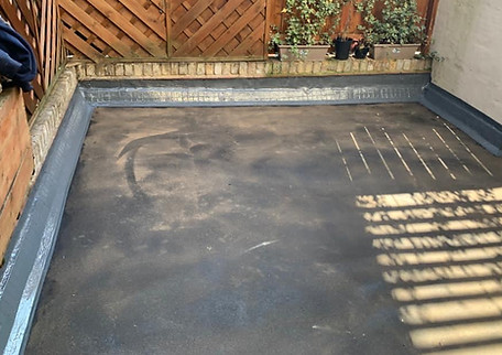 The image depicts a flat, solved roof repair backyard patio that appears to be empty and treated with a dark coating, giving it a shiny look. Shadows from the wooden fence are cast on the ground, adding depth to the scene.