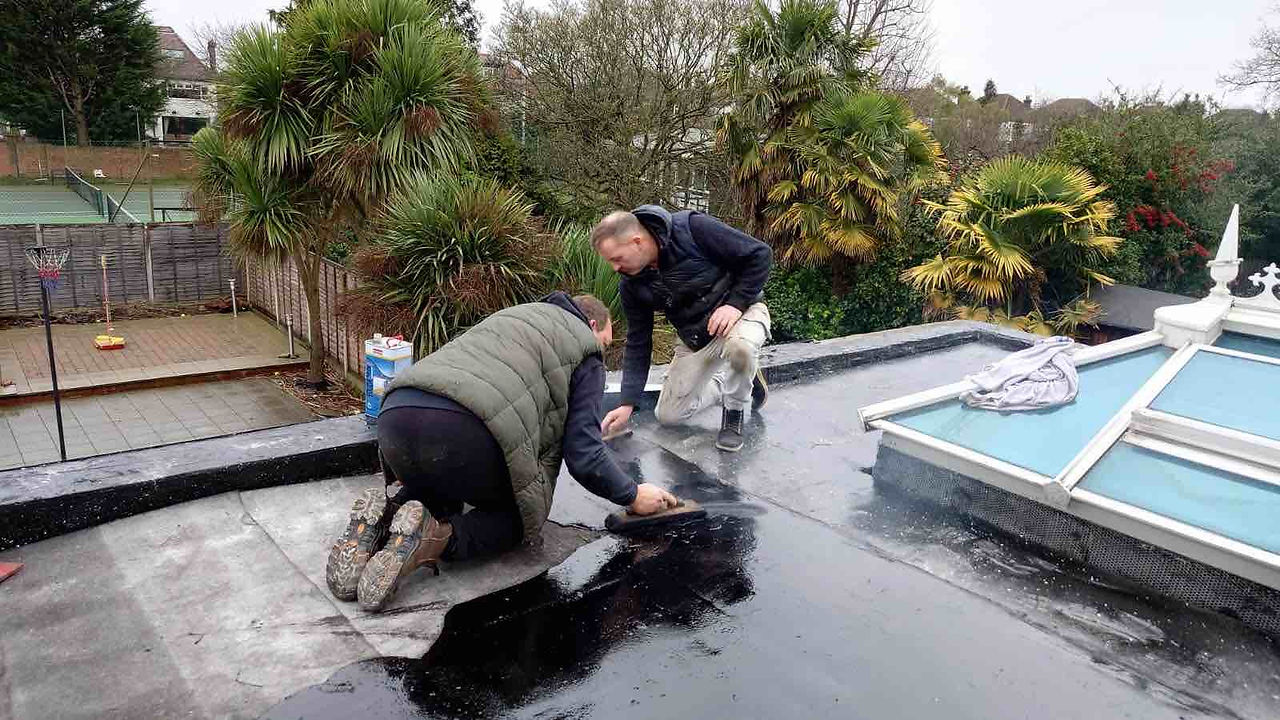 Two workers repairing a flat roof near a skylight with a lush garden view.
