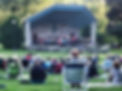 A photo of a live performance at the half shell located at Wood Park on Park St in Hudson - performers are on the stage and guests are sitting on the lawn watching