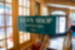 A green sign reading Men's Shop Downstairs inside of a clothing store.