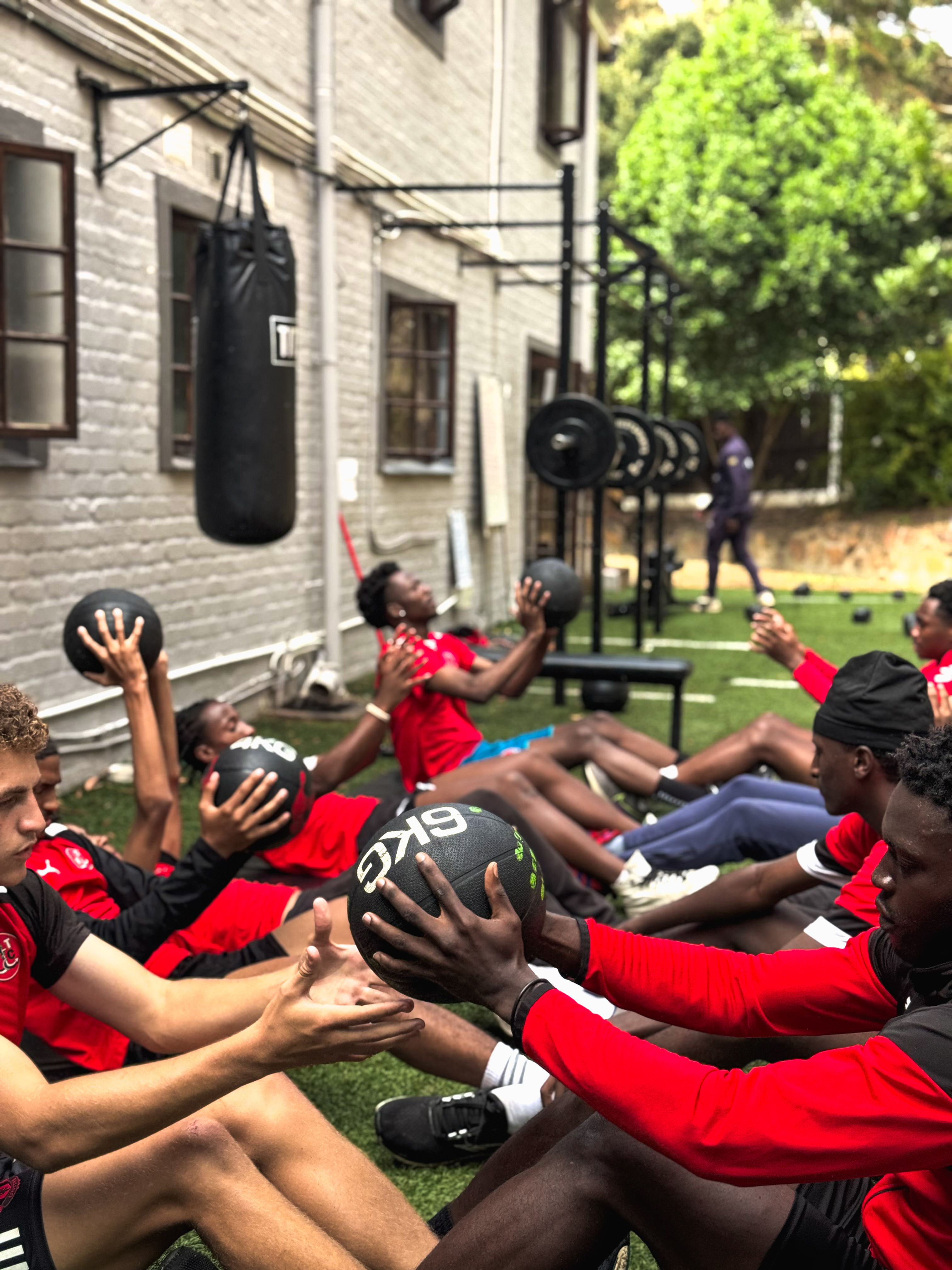  Players from Fleetwood Town International Football Academy training on a sandy beach under a bright blue sky. They are wearing red shirts and blue shorts, engaging in a beach soccer drill with an airborne ball. Some players are barefoot, while others stand ready to receive the ball. The ocean waves roll in the background, with a red-and-yellow flag marking a designated area on the shore. The session reflects the academy’s dynamic and adaptive training approach, combining skill development with a fun, challenging beach environment.