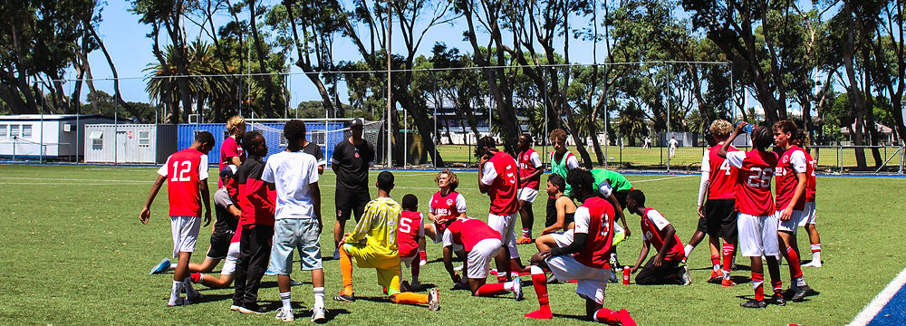 Players from Fleetwood Town International Football Academy training on a sandy beach under a bright blue sky. They are wearing red shirts and blue shorts, engaging in a beach soccer drill with an airborne ball. Some players are barefoot, while others stand ready to receive the ball. The ocean waves roll in the background, with a red-and-yellow flag marking a designated area on the shore. The session reflects the academy’s dynamic and adaptive training approach, combining skill development with a fun, challenging beach environment.