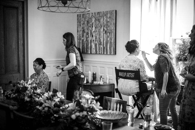 A bride sits getting her makeup done while facing the large windows in the dining room at Boxwood Villa while her bridal party also gets ready behind her.