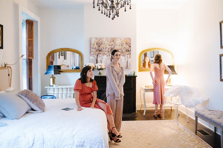 The bride and her mother look expectantly at the doorway while a bridesmaid looks at herself in the mirror in the master suite bedroom on the first floor of Boxwood Villa.