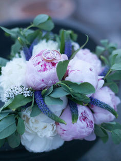 Peonies, white roses and a small purple flower create a stunning bouquet displaying the couple's rings on top.