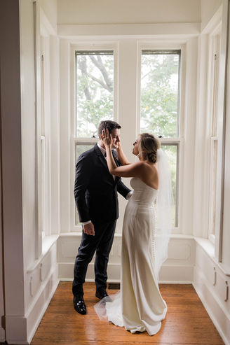 A bride adoringly cups her groom's face in her hands after their first look moment in front of the beautiful windows of Boxwood Villa.
