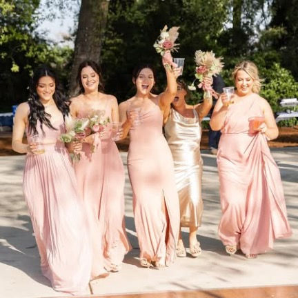 Five women in pink dresses smiling and cheering outdoors