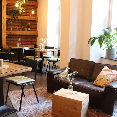 Ein Gastraum in einem Restaurant mit einer dunklen Zweisitzer-Couch, schwarzen Stühlen mit hellgrünen Sitzkissen und Holztischen.