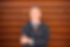 Eric Sullivan in navy suit and brown striped tie standing against wooden background with arms crossed and smiling