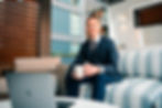 Eric Sullivan wearing navy suit and brown striped tie sitting on couch in outdoor working space holding a coffee cup and smiling