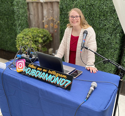 dj b diamond wearing a red dress with a cream colored cardigan standing at her dj mixing table with her equipment laptop and microphone with a green plant wall background outside