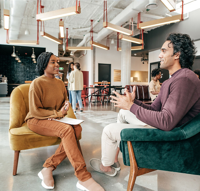 Two young professionals sitting together in a modern office environment while discussing collaboration and community health initiatives.
