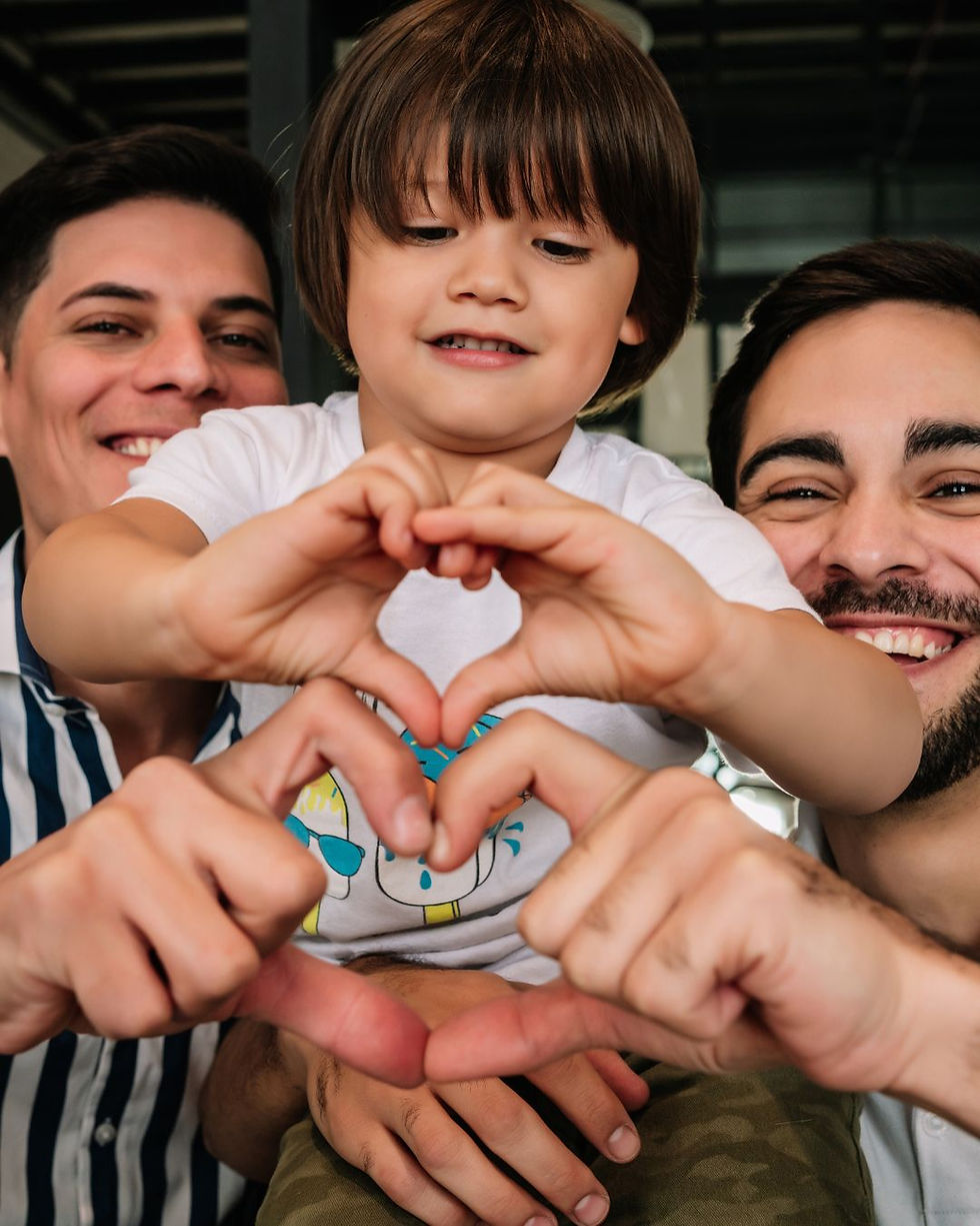 Two fathers and their son smiling together and making a heart shape with their hands to represent family, support, and community healthcare.