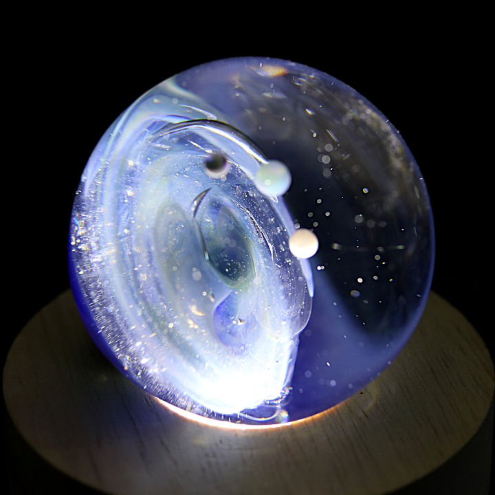 Thumbnail: Veilleuse Galaxie avec une voie lactée de couleur bleu et trois planètes en orbite. La Galaxie est posée sur un socle en bois lumineux. Vue de la Galaxie d'un autre angle qui nous permet de voir le vortex galactique