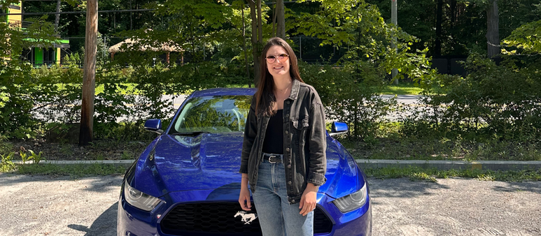 Photo de Manon Pumilia devant une voiture de location Mustang bleue via l’application Turo, prise au Québec, illustrant un road trip au Canada.