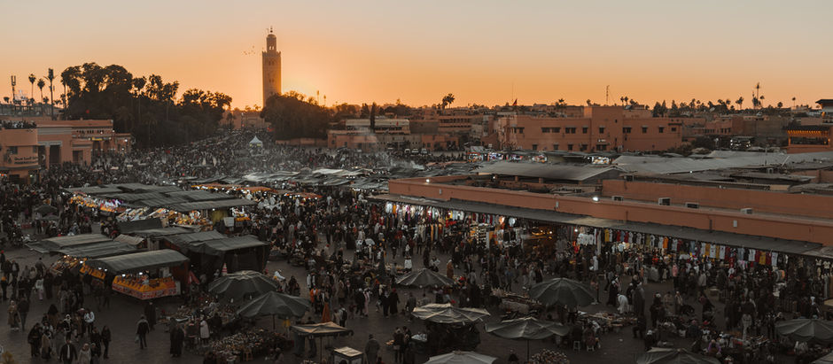 Djemaa el Fna in Marrakesch bei Sonnenuntergang. Viele Menschen und Verkaufsstände. Orangefarbener Himmel im Hintergrund, lebhafte Stimmung.