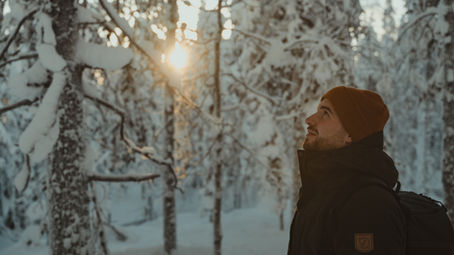 Mann im Winterwald in Finnisch Lappland schaut lächelnd zur Sonne. Hintergrund verschneite Bäume, warme Farben. Trägt braune Mütze und schwarze Jacke.