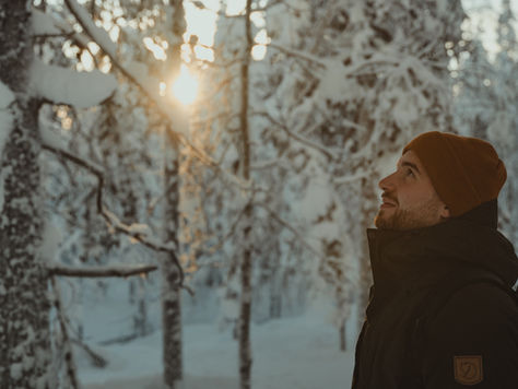 Mann im Winterwald in Finnisch Lappland schaut lächelnd zur Sonne. Hintergrund verschneite Bäume, warme Farben. Trägt braune Mütze und schwarze Jacke.