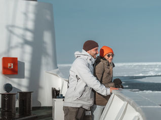 Zwei Personen in Winterkleidung stehen lächelnd auf einem Expeditionsschiffs, umgeben von Eis und Meer in der Arktis. Sonne scheint, klare blaue Himmel.