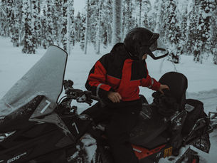 Person in roter Jacke auf Schneemobil im verschneiten Wald in Finnisch Lappland, hält Helm, winterliche Atmosphäre, Schnee bedeckt Bäume im Hintergrund.