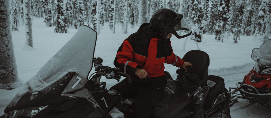 Person in roter Jacke auf Schneemobil im verschneiten Wald in Finnisch Lappland, hält Helm, winterliche Atmosphäre, Schnee bedeckt Bäume im Hintergrund.