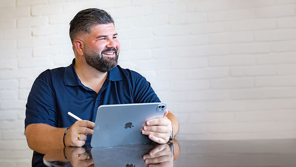 Inhaber hält Tablet in der Hand, sitzt am Tisch und lacht.