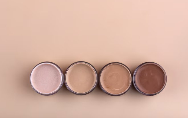 Four round containers of foundation in shades from light to dark on a beige background, showcasing makeup diversity.