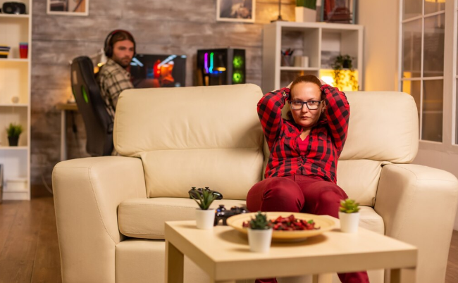 Woman sitting on a beige sofa in a red plaid shirt, arms raised, looking tense. Man gaming in the background. Cozy, lit living room.