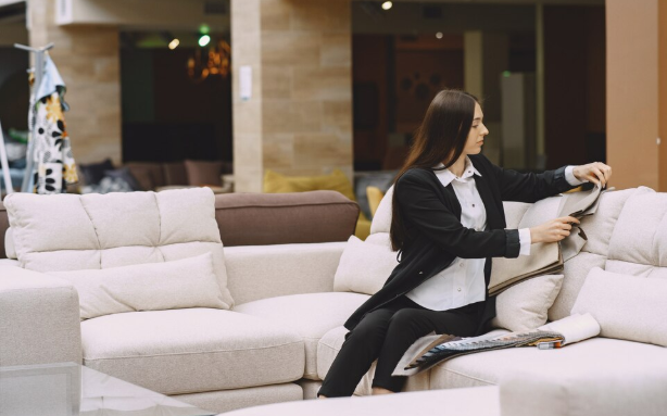 Woman in a black suit sits on a beige sofa, reading a magazine in a modern showroom. Neutral colors and relaxed ambiance.