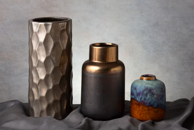 Three decorative vases on a draped gray fabric. Textured silver, bronze metallic, and a small blue-orange vase, set against a neutral backdrop.