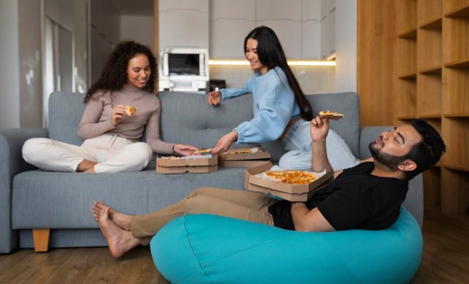 Three people enjoy pizza in a cozy living room. Two women sit on a gray couch, smiling, while a man lounges on a blue beanbag.