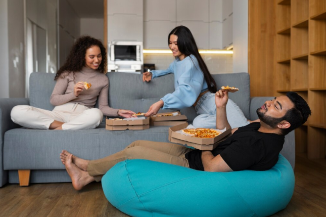 Three people enjoy pizza in a cozy room. One reclines on a blue bean bag, while the others sit on a gray sofa, surrounded by pizza boxes.