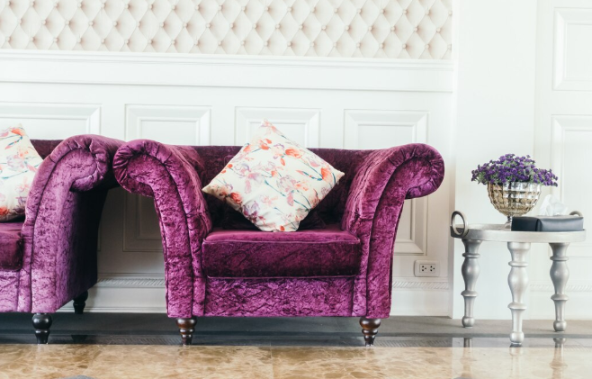 Purple velvet armchair with floral pillow in elegant room. Marble floor, white paneled walls, and silver table with purple flowers. Luxurious mood.
