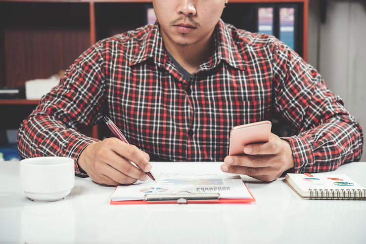 Man in plaid shirt writes on clipboard while holding a phone. Notebook and coffee cup on white table. Office shelves in background.